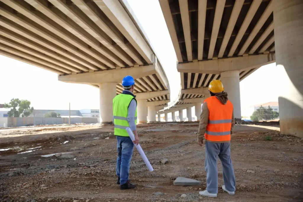 Two workers in hi-vis and hard hats inspecting the underside of a concrete bridge deck on a civil site